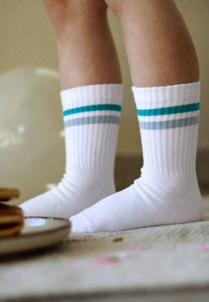 Beine mit weißen Crew-Socken mit türkis- und grauen Streifen, die auf einem Teppich neben einem Teller mit gestapelten Pfannkuchen stehen.