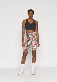Sujetador deportivo negro con tirantes ajustables, combinado con shorts de bicicleta con estampado floral colorido. La modelo lleva zapatillas deportivas blancas y calcetines.
