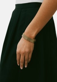 Kullast metallist käevõru punutud disainiga, mis on käsivarrel. Taustal on tumeroheline seelik. Sile tekstuur läikiva viimistlusega.