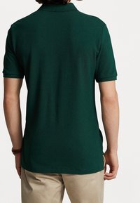 Ciemnozielona koszulka polo z klasycznym kołnierzykiem, krótkimi rękawami i bocznymi rozcięciami, wykonana z fakturowanej tkaniny, noszona na jasnych spodniach.