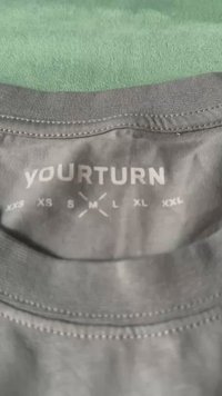 Sivá tkanina s jemnou textúrou, so štandardným okrúhlym výstrihom a potlačenou etiketu, na ktorej sú veľkosti od XXS po XXL v bielej farbe.