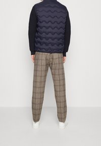 Marinblå pufferväst med vågig textur, kombinerad med lösa, grå rutiga byxor med en checkmönster. Vita sneakers fulländar outfiten.
