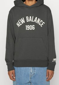 Felpa con cappuccio grigia con tasca a marsupio frontale, testo curvo "NEW BALANCE" e "1906" in crema, polsini a costine e dettaglio del logo sulla manica.