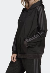 Svart huvtröja i texturerat tyg med en känguruficka och tre vertikala ränder i en mörkare nyans på ärmarna.