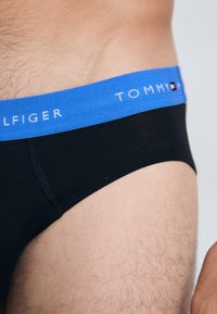 Schwarze Baumwollslips mit einem blauen Elastikbund, der mit weißem "TOMMY HILFIGER"-Text und Logodetails versehen ist. Glatte Textur, körpernahe Passform.