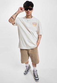 Christ-farvet oversized t-shirt med frontgrafik, beige corduroy shorts og grå sneakers med hvide accenter og mørke snørebånd.