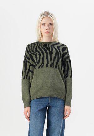 Pull en maille vert avec des rayures noires de zèbre, col rond et épaules tombantes. Texture douce, coupe décontractée. Associé à un jean en denim bleu.