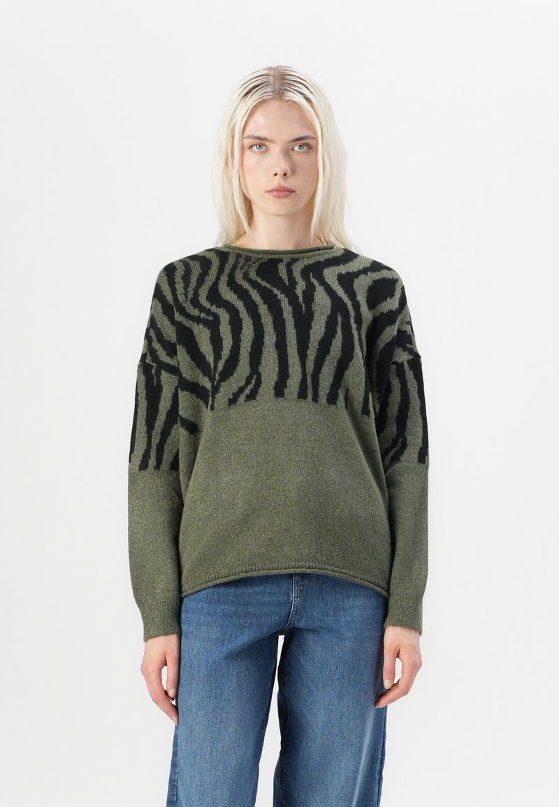 Grüner Strickpullover mit schwarzen Zebra-Streifen, Rundhalsausschnitt und überschnittenen Schultern. Weiche Textur, lockere Passform. Kombiniert mit blauen Jeans.