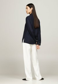 Camicia a manica lunga blu navy con colletto, spacco laterale e bottoni dorati. Abbinata a pantaloni bianchi comodi e scarpe nere.