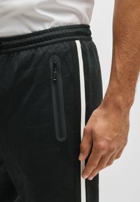 Svarta träningsbyxor med en texturerad väv, sidoficka med dragkedja och vit sidolinje. Logotypen syns på nederdelen av benet.