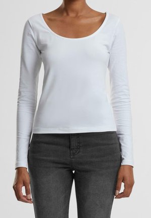 Kobieta w dopasowanej białej koszulce z długim rękawem i dekoltem w serek oraz w ciemnoszarych, wysokim stanem jeansach, stojąca na tle jednolitego tła.