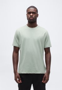 Άνδρας που στέκεται προσανατολισμένος προς τα εμπρός, φοράει ανοιχτό πράσινο κοντομάνικο μπλουζάκι και μαύρο παντελόνι, σε ένα απλό λευκό φόντο.