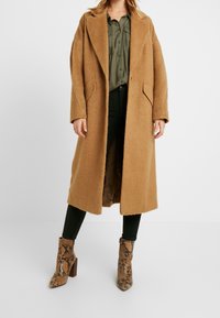 Cappotto in lana color tan con vestibilità rilassata, ampi revers e tasche laterali. Indossato sopra una camicia verde oliva, abbinato a stivaletti alla caviglia con stampa serpente.
