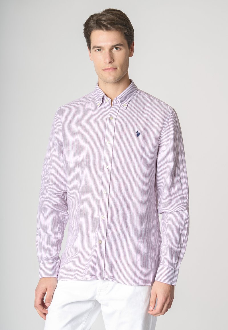 Un giovane indossa una camicia button-up a righe viola e bianche con colletto, in piedi contro uno sfondo neutro. Mani in tasca.