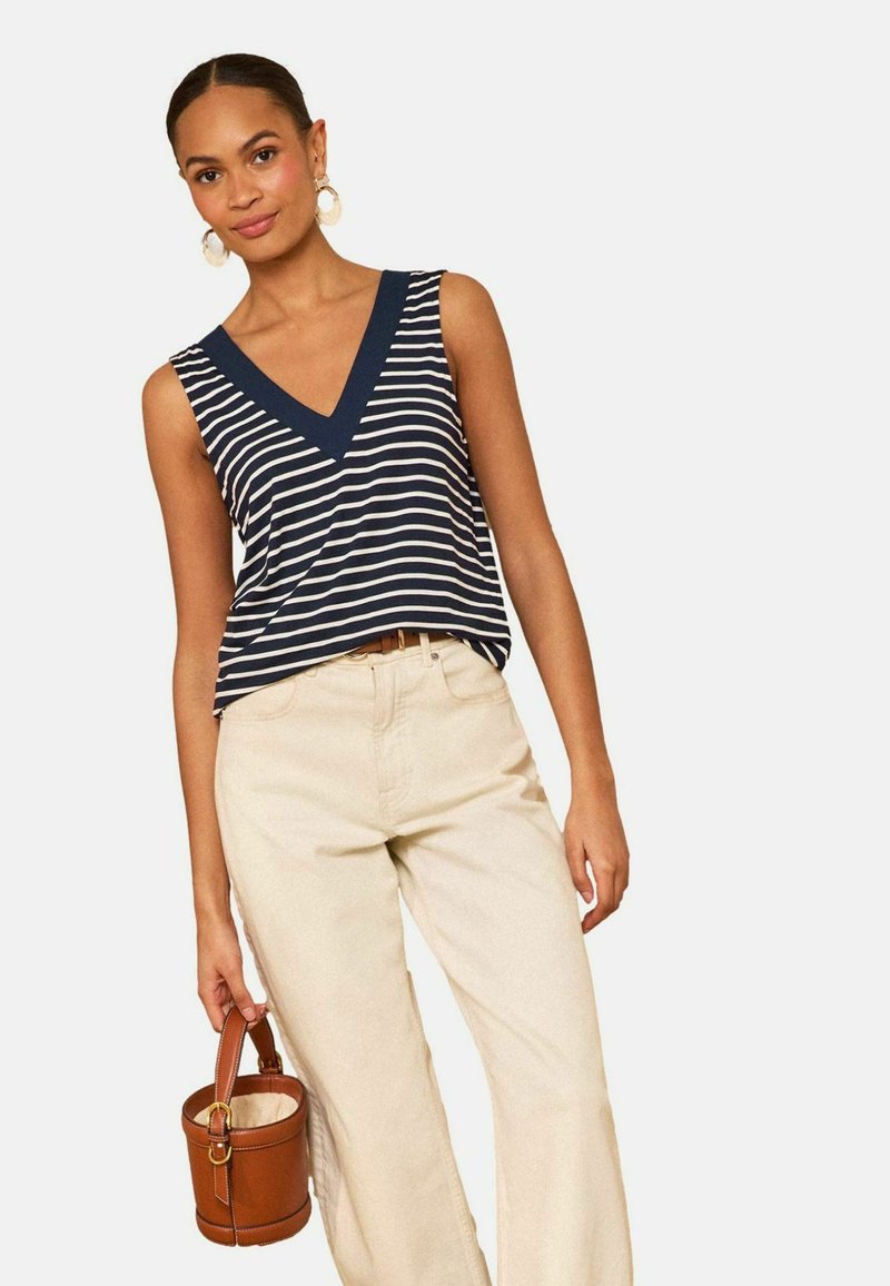 Femeie purtând un top fără mâneci cu dungi albastre și albe și pantaloni crem, ținând o geantă mică maro din piele, de tip găleată, de mânere.