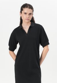 Black polo dress with short sleeves, featuring a collared neck and front placket. Smooth texture, knee-length with a relaxed fit.