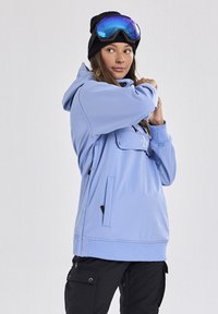 Hellblaue Pullover-Hoodie mit einer Front-Reißverschlusstasche, verstellbarem Kordelzug, seitlichen Reißverschlusstaschen, kombiniert mit schwarzen Hosen und einer Schutzbrille.