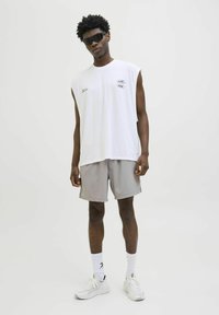 Camisa sin mangas de algodón blanco con detalles impresos, combinada con pantalones cortos gris claro. El modelo lleva gafas de sol negras y zapatillas blancas.