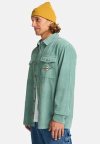 Green corduroy shirt with two chest pockets, button closure, and an embroidered logo. Paired with a yellow beanie. Casual fit and texture.