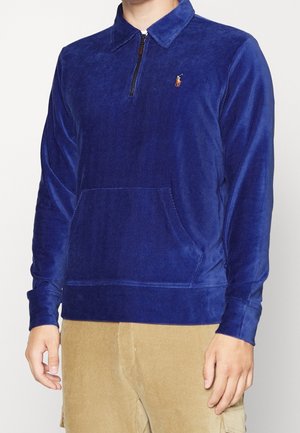 Homme portant un pull en velours côtelé bleu avec une demi-fermeture éclair et une poche kangourou, associé à un pantalon en velours côtelé beige.