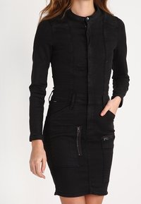 Czarna sukienka dżinsowa z wysokim kołnierzem, długimi rękawami, dopasowanym krojem i wieloma zamkami błyskawicznymi z przodu.