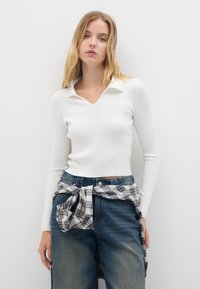 Top bianco a maniche lunghe con collo a V, abbinato a jeans blu a gamba larga e una camicia a quadri annodata in vita.