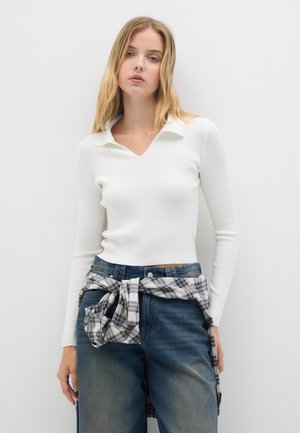 Top bianco a maniche lunghe con collo a V, abbinato a jeans blu a gamba larga e una camicia a quadri annodata in vita.