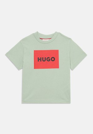 Svijetlozeleni pamučni majica s kratkim rukavima sa crvenim kvadratom na kojem je crni tekst "HUGO" centriran na prednjoj strani. Ima običan vratni izrez.