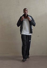 Mand i sort-hvid Adidas tracksuit justerer jakkekraven, mens han står mod en simpel betonvæg i et indendørs miljø.