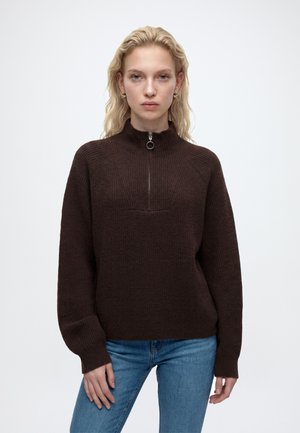 Femme aux cheveux blonds portant un pull côtelé marron foncé avec une demi-fermeture éclair et un jean bleu, debout devant un fond blanc, face à l'avant.