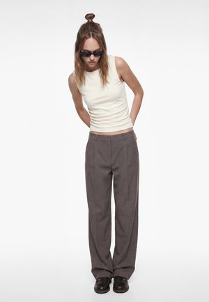 Jeune femme portant un haut blanc sans manches, un pantalon large gris, des mocassins noirs et des lunettes de soleil noires, debout les mains derrière le dos.