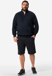 Námořnická mikina na zip s přední kapsou, spárovaná s tmavými šortkami a černými teniskami, stojící na jednobarevném pozadí.