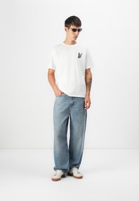 White cotton t-shirt with a butterfly graphic, paired with light blue wide-leg jeans and white sneakers with brown accents. Gray sunglasses.