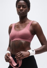 Αθλητικό άτομο με ροζ αθλητικό σουτιέν και μαύρο κολάν, φορώντας λευκά περιβραχιόνια, κρατώντας μια ροζ λωρίδα αντίστασης, στέκεται σε απλό φόντο.