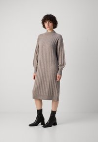 Virkattu mekko vaaleanruskeana, jossa on pystysuora ribbitekstuuri, korkea kaulus ja pitkät puffihihat, yhdistettynä mustiin nilkkabootseihin.