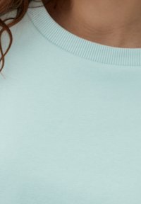 Cuello y parte superior del pecho de una persona que lleva un suéter de cuello redondo de canalé azul claro, con cabello rizado castaño a los lados.