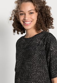 T-shirt preta com mangas curtas, apresentando um padrão salpicado em bege claro. Textura suave, decote redondo, ajuste descontraído.