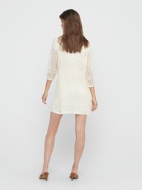 Woman with long brown hair wearing a white crochet cardigan, white shorts, and brown heeled sandals, standing facing away on a plain background.