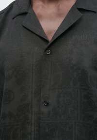 Moški, ki nosi črno srajco z nežnim reliefnim cvetličnim vzorcem, odprtim ovratnikom in dvema sprednjima črnima gumboma, pritrjenima.