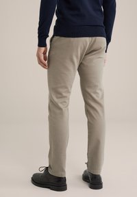 Chino bej cu o croială slim, având o textură netedă. Poartă pantofi negri cu șireturi. Pulover bleu închis parțial vizibil.