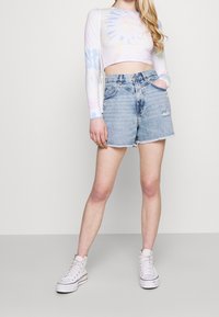 Top corta de manga larga con efecto tie-dye claro, combinado con shorts de mezclilla de cintura alta y desgastados, con bordes deshilachados. Se lleva con zapatillas blancas de caña alta.