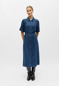 Femme portant une robe en denim bleu mi-longue avec une broderie florale sur le col et des bottes noires montantes, debout les mains derrière le dos.