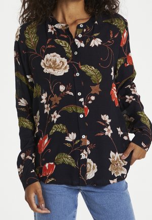 Woman wearing a black button-up blouse with a floral pattern in white, green, and red, paired with blue jeans, hands in pockets.