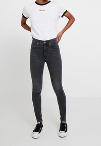 Jeans negros ajustados de cintura alta, con un diseño de cinco bolsillos y costuras mínimas. Combinados con una camiseta blanca con un logo rojo.