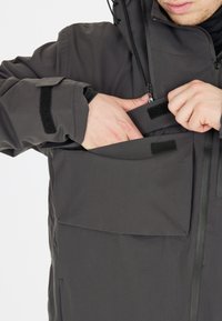 Tummanharmaa takki, jossa on vetoketjulukko ja näkyvät tarranauhakäät. Malli käyttää etutaskua, jossa on tarranauhakiinnitys.