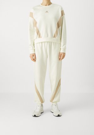 Osoba ubrana w kremowo-beżowy dres Adidas z pasującymi sneakersami, stojąca na jednolitym białym tle.