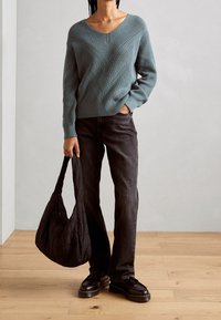 Morski zielony sweter na drutach z dekoltem w serek i wzorem w jodełkę, zestawiony z czarnymi jeansami o prostych nogawkach oraz masywnymi czarnymi butami. Trzyma pikowaną czarną torbę.