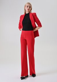 Abito rosso su misura con maniche lunghe e pantaloni a gamba larga, abbinato a una maglia nera aderente e testurizzata e ballerine nere con fiocco.