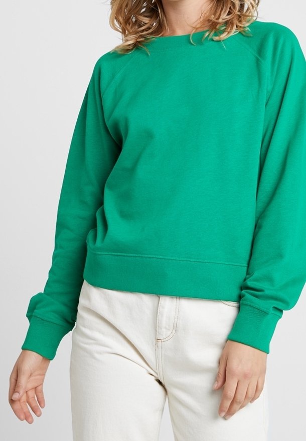 Femme portant un sweat-shirt vert à manches longues et un pantalon blanc, debout devant un fond clair uni.