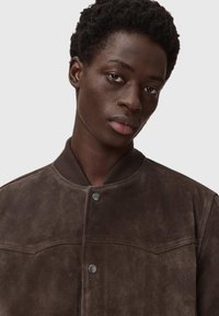 Brązowa skórzana kurtka bombowa z okrągłym kołnierzem, zapięciem na guziki i delikatnymi detalami szycia. Gładka faktura, nowoczesny krój, minimalistyczny design.